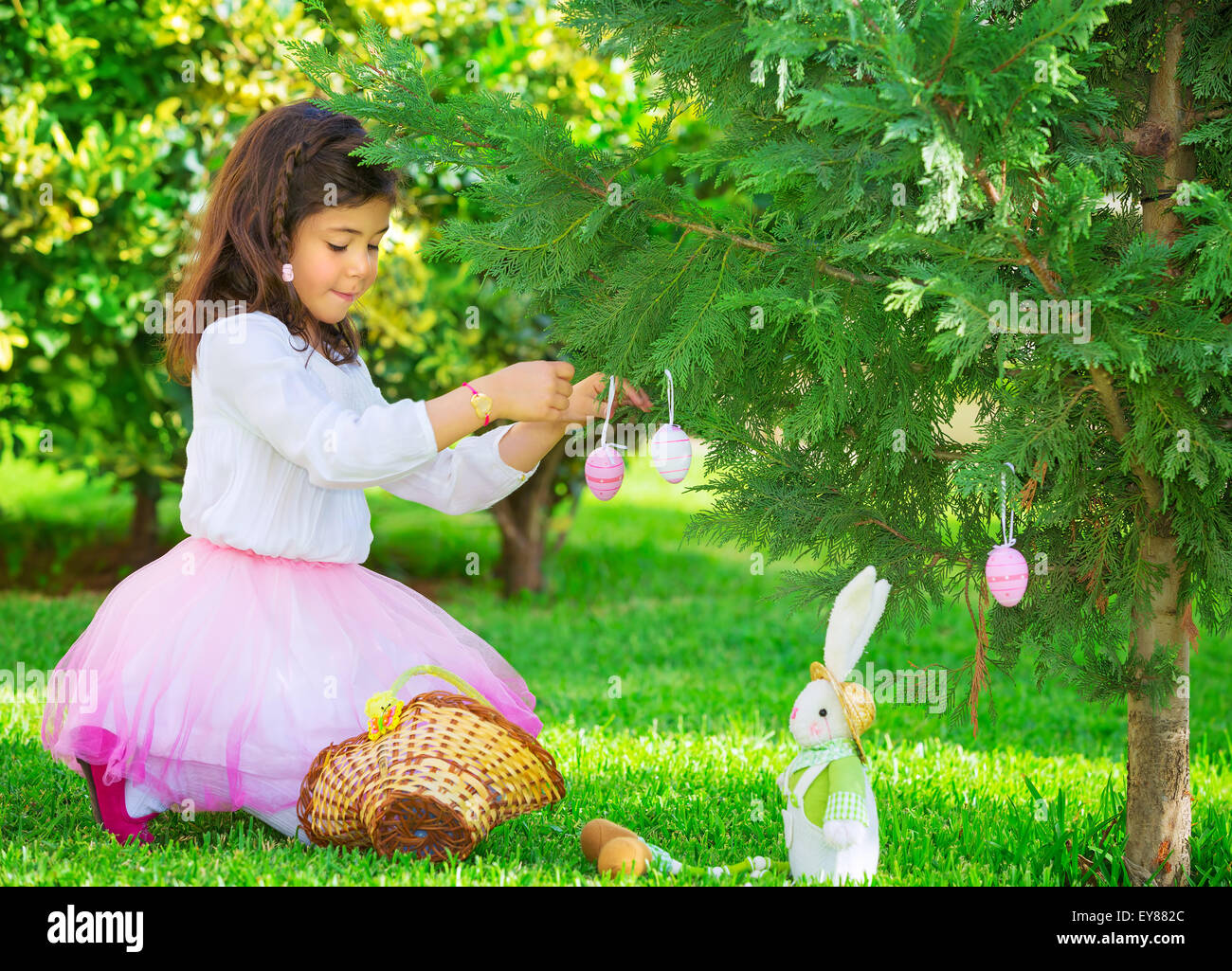 Adorable little girl having fun outdoors with Easter bunny toy, decorated fresh green tree with coloring eggs, Happy Easter Stock Photo