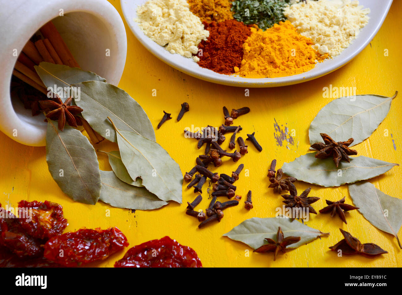 Colorful cooking spices and herbs on a round plate with cinnamon sticks, bay leaves and star anise in mortar and pestle on yello Stock Photo
