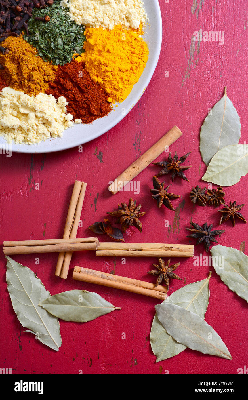 Colorful cooking spices and herbs on round plate with cinnamon sticks, star anise and bay leaves on vintage red wood table. Stock Photo
