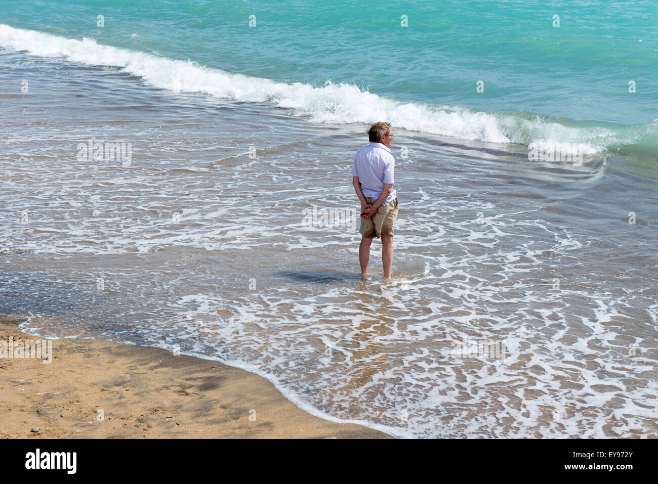 middle aged man wearing shorts and summer shirt standing ankle deep in the surf on a sandy beach at freshwater isle of wight Stock Photo