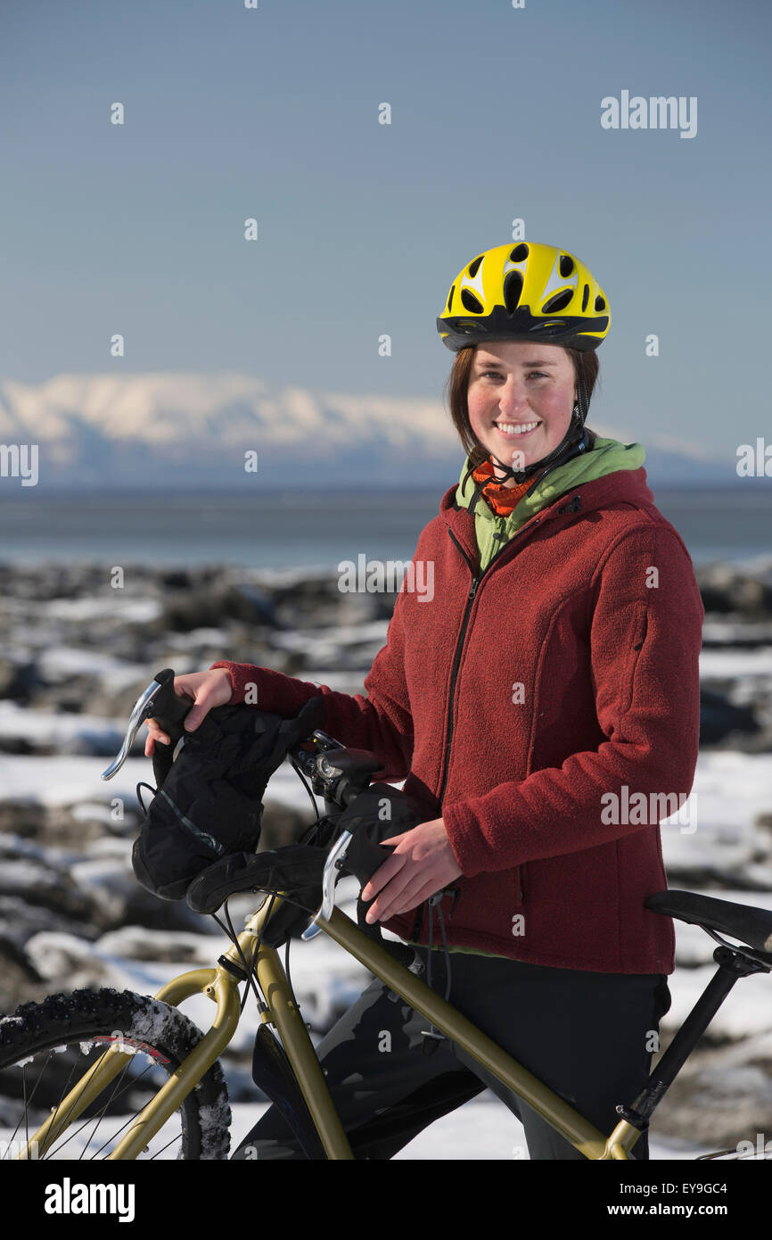 A young woman stands with her bicycle on ice next to the Tony Knowles Coastal Trail, Anchorage, Southcentral Alaska, USA. Stock Photo