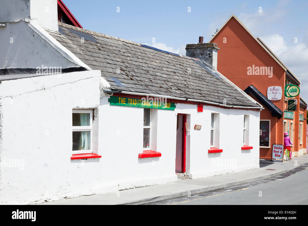 A Woman On A Rented Bicycle Rides By Shops Along A Street; Doolin, County Clare, Ireland Stock Photo