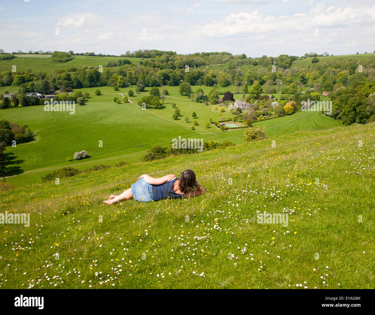 Woman lying on chalk grassland looking into deep green valley containing Rainscombe House, Oare Hill, Wiltshire, England, UK Stock Photo