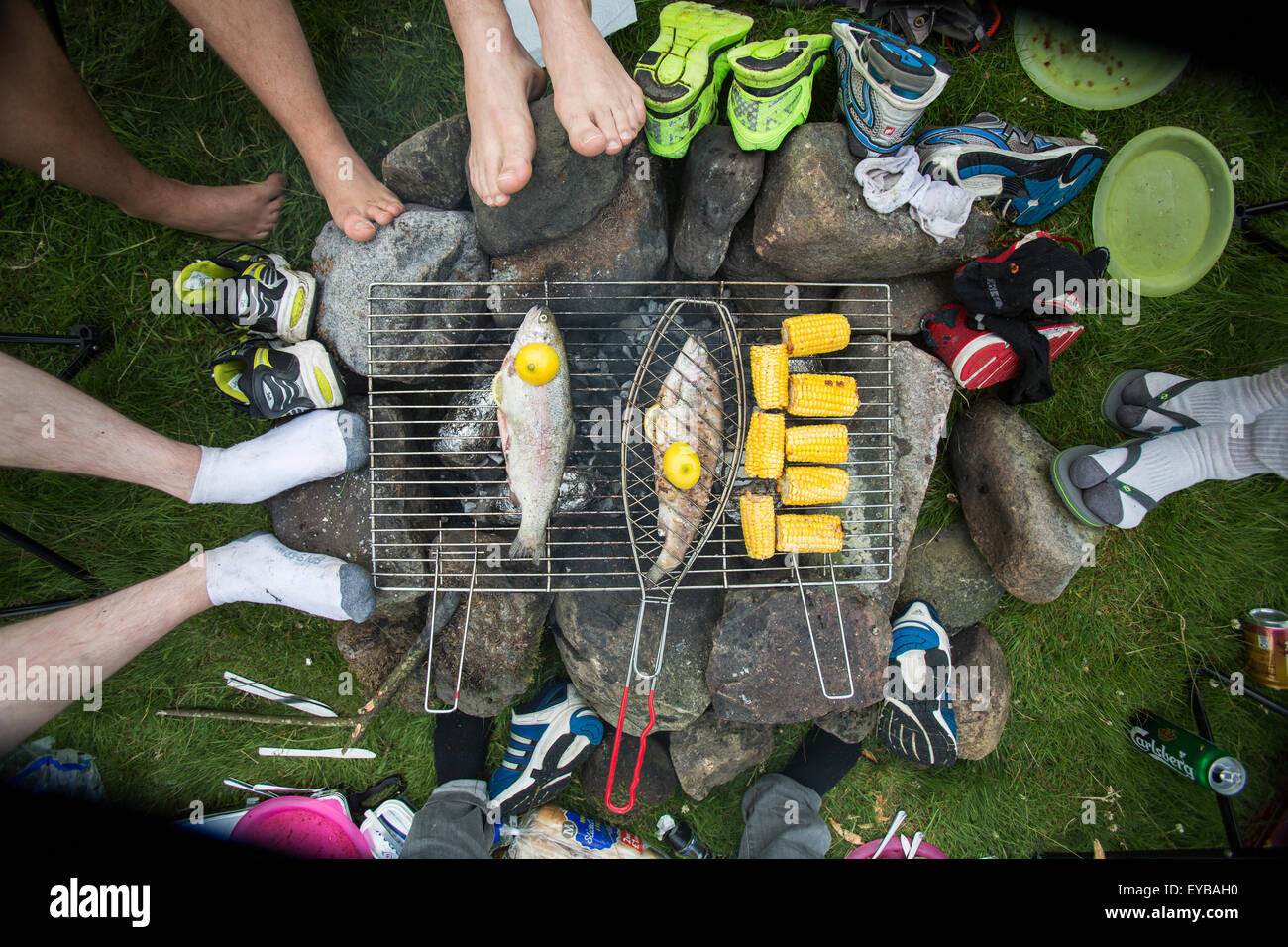 friends sit around a camp fire cooking some fish and corn on the cob in the hills of Scotland, 2015. Stock Photo