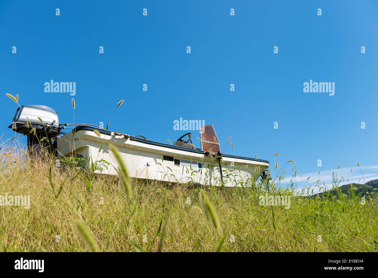 An old abandoned boat surrounded by overgrown grass with a clear blue sky, small clouds and mountains in the distant background. Stock Photo