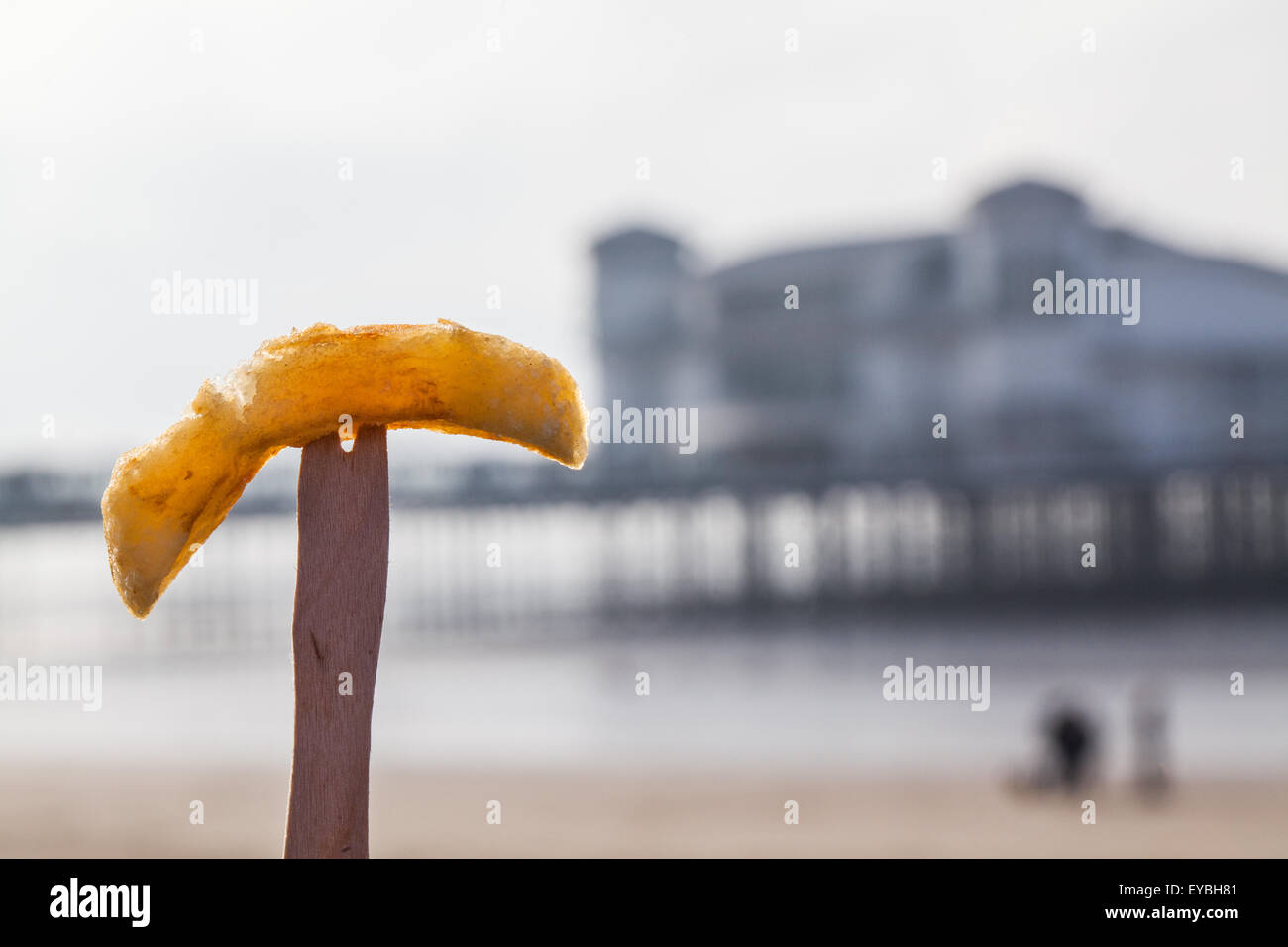 A freshly fried British chip presented on a traditional wood chip shop fork at the seaside with a blurred pier in the background Stock Photo