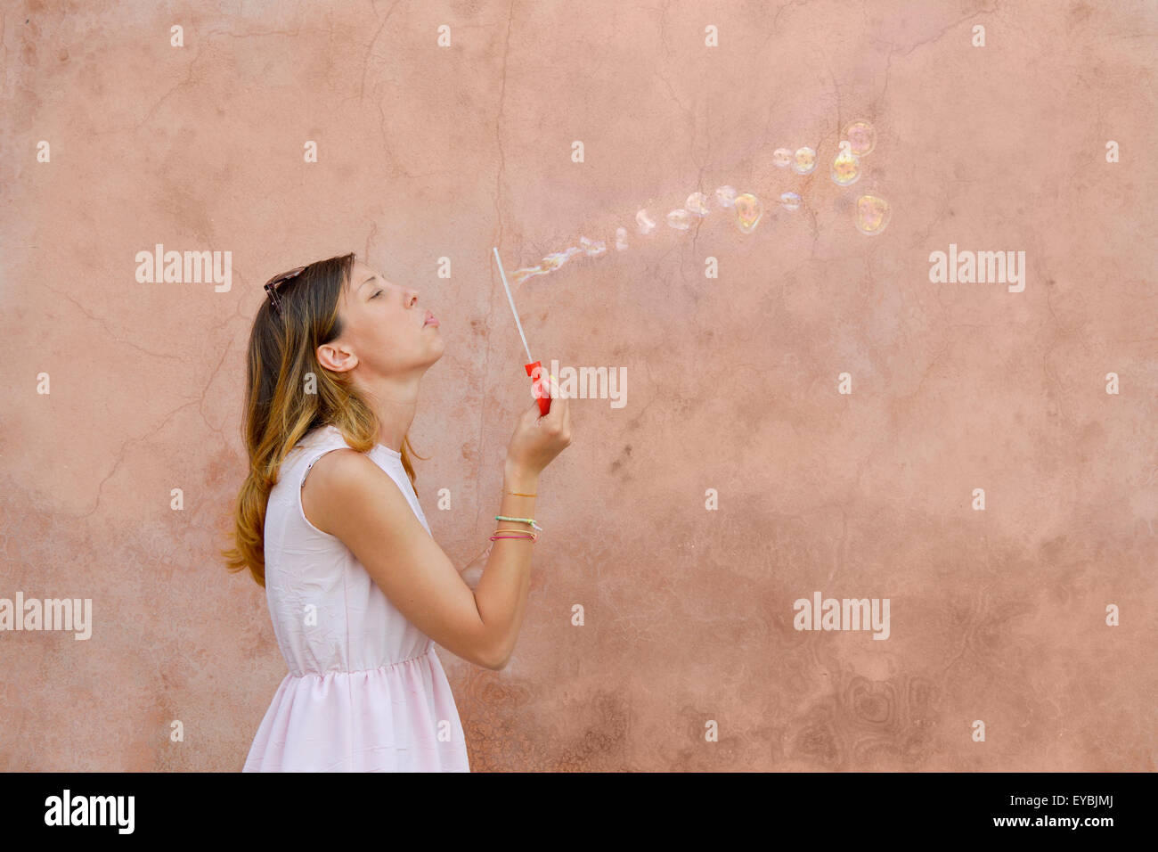 Girl blowing soap bubbles against colourful backdrop wearing pink dress Stock Photo