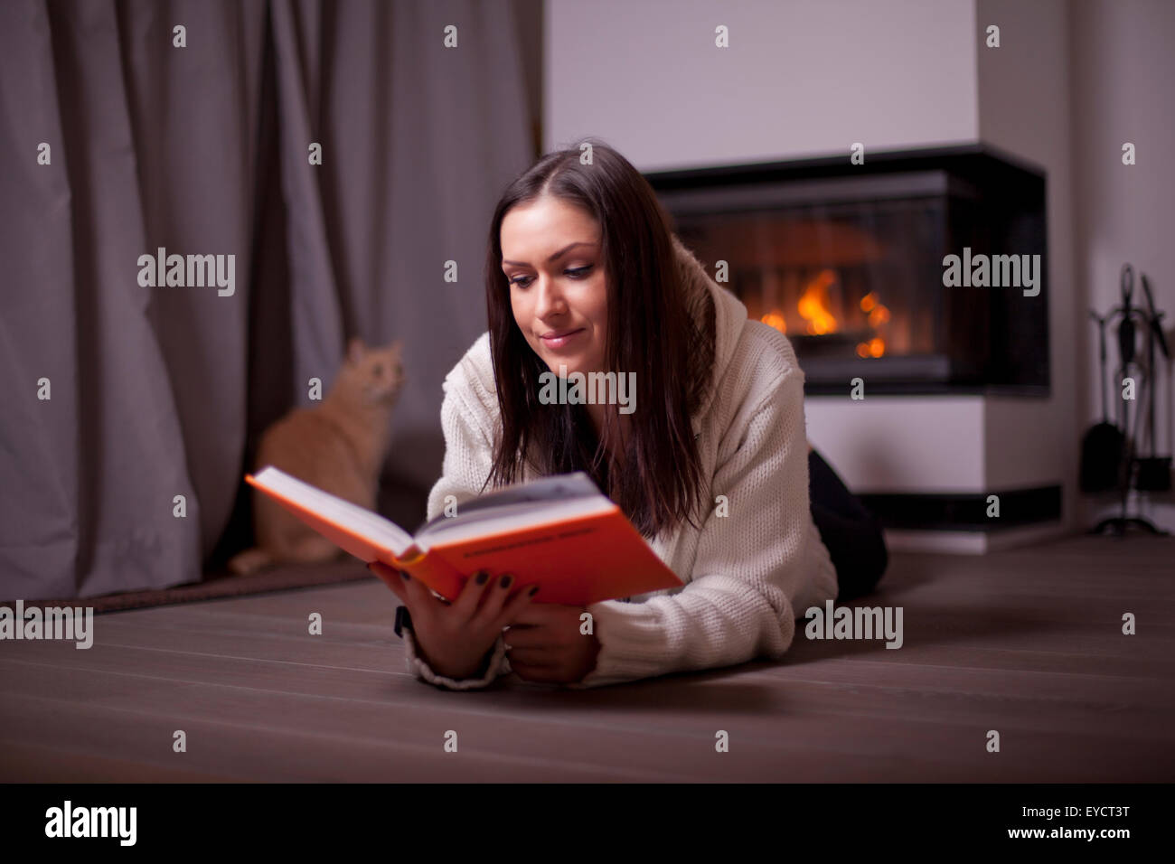 Beautiful woman lying on floor and reading book by fireplace and cat behind Stock Photo