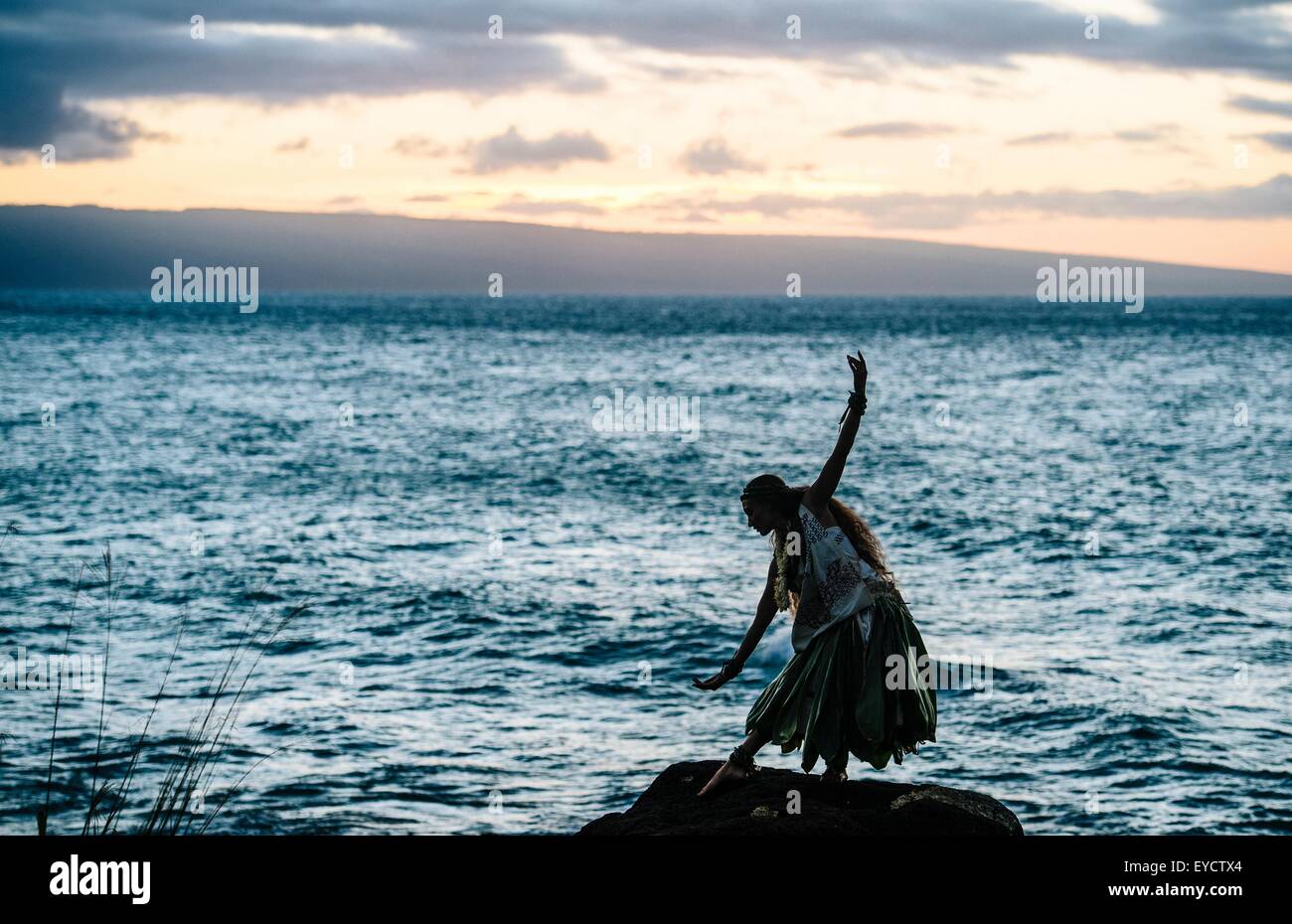 Silhouetted woman hula dancing on coastal rocks wearing traditional costume at dusk, Maui, Hawaii, USA Stock Photo