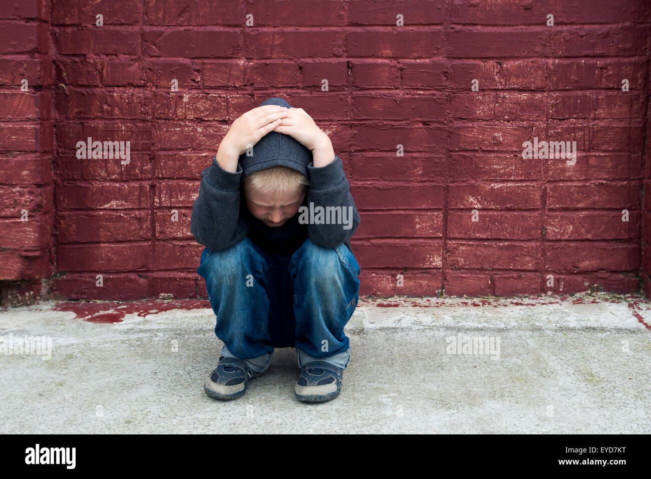 Depressed abused upset crying child (boy, kid, teen) sitting near red brick wall Stock Photo