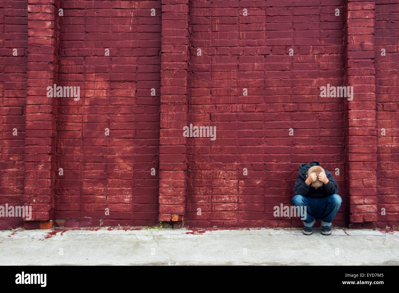 Depressed abused upset crying child (boy, kid, teen) sitting near red brick wall Stock Photo
