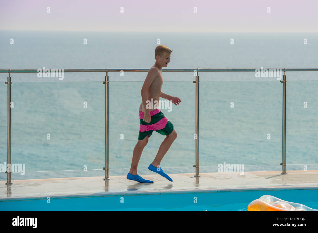 A boy wearing flippers walks by a swimming pool with sea behind hime