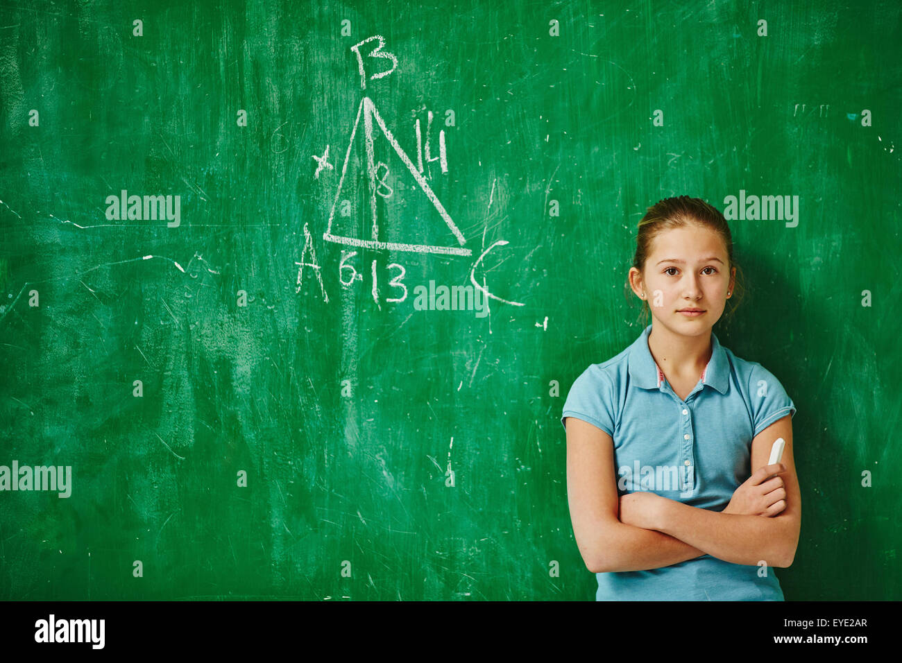 Cute schoolgirl looking at camera while standing by blackboard with geometric task on it Stock Photo