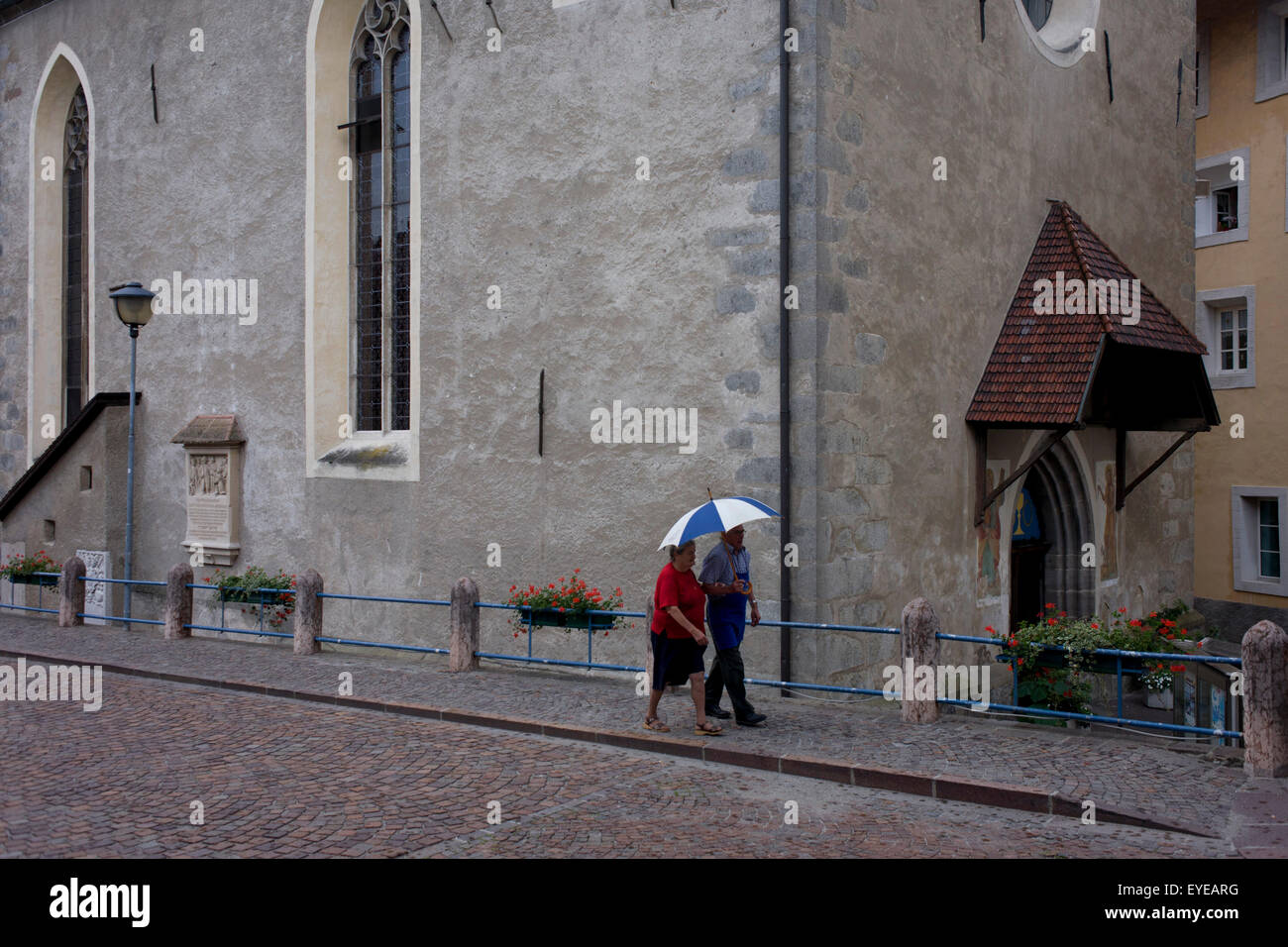 Elderly couple walk in the rain past the church in Klausen-Chiusa in the Italian south Tyrol. Stock Photo