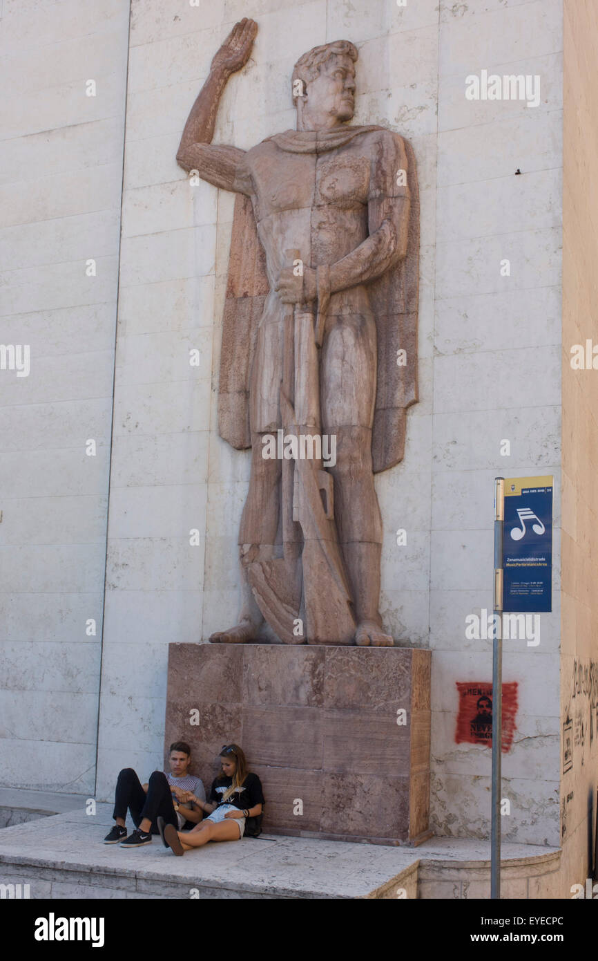 Young couple site beneath Fascist statue outside a government building in the northern Italian regional city of Trento. Stock Photo