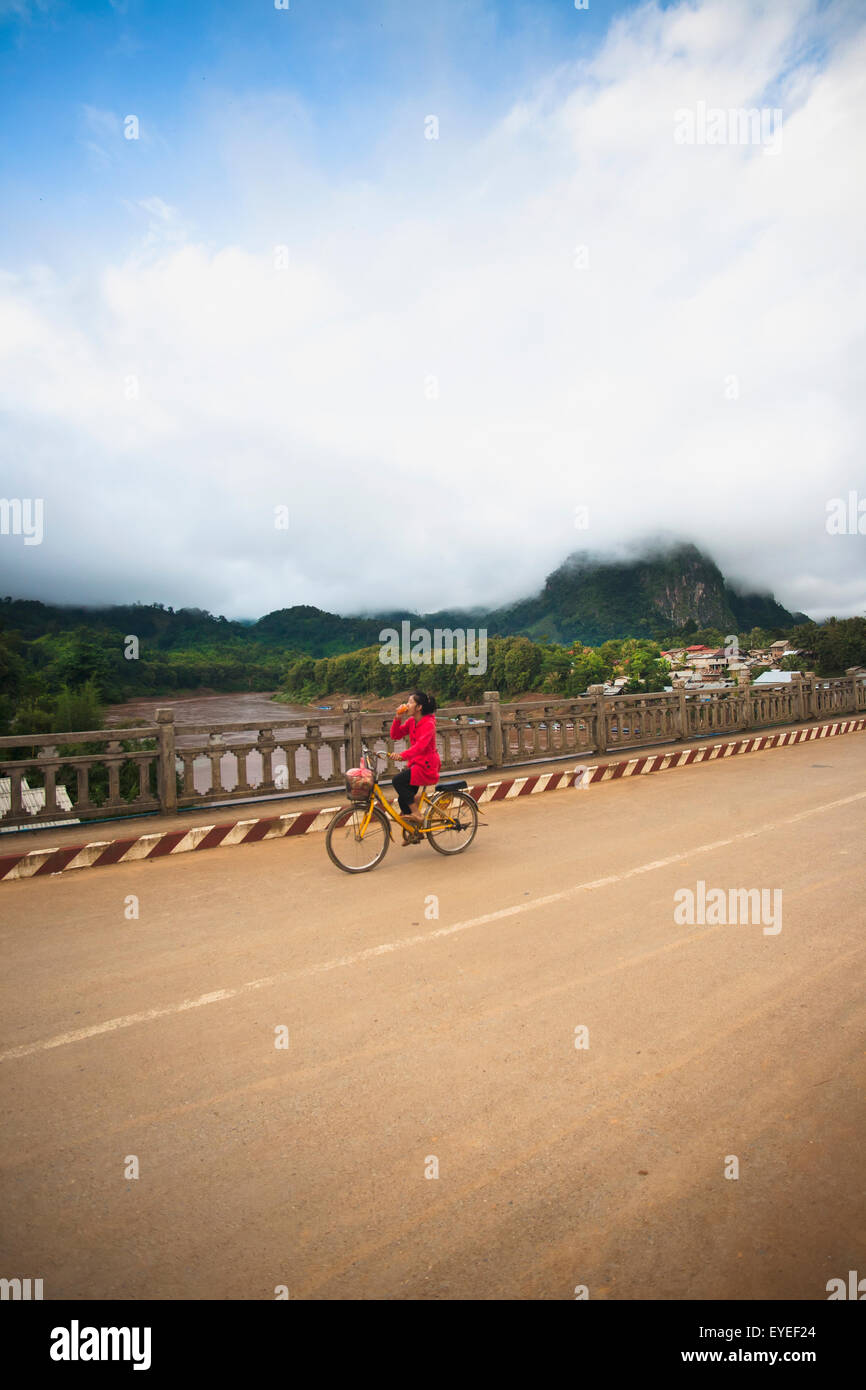 A young woman cycles in Northern Laos town of Nong Khiaw, along the Ou River; Nong Khiaw, Laos Stock Photo