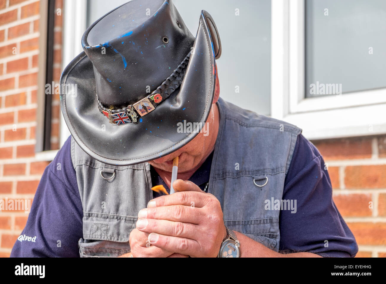 A male Caucasian lighting a cigarette with his hands cupped around the cigarette wearing a leather cowboy hat. Stock Photo