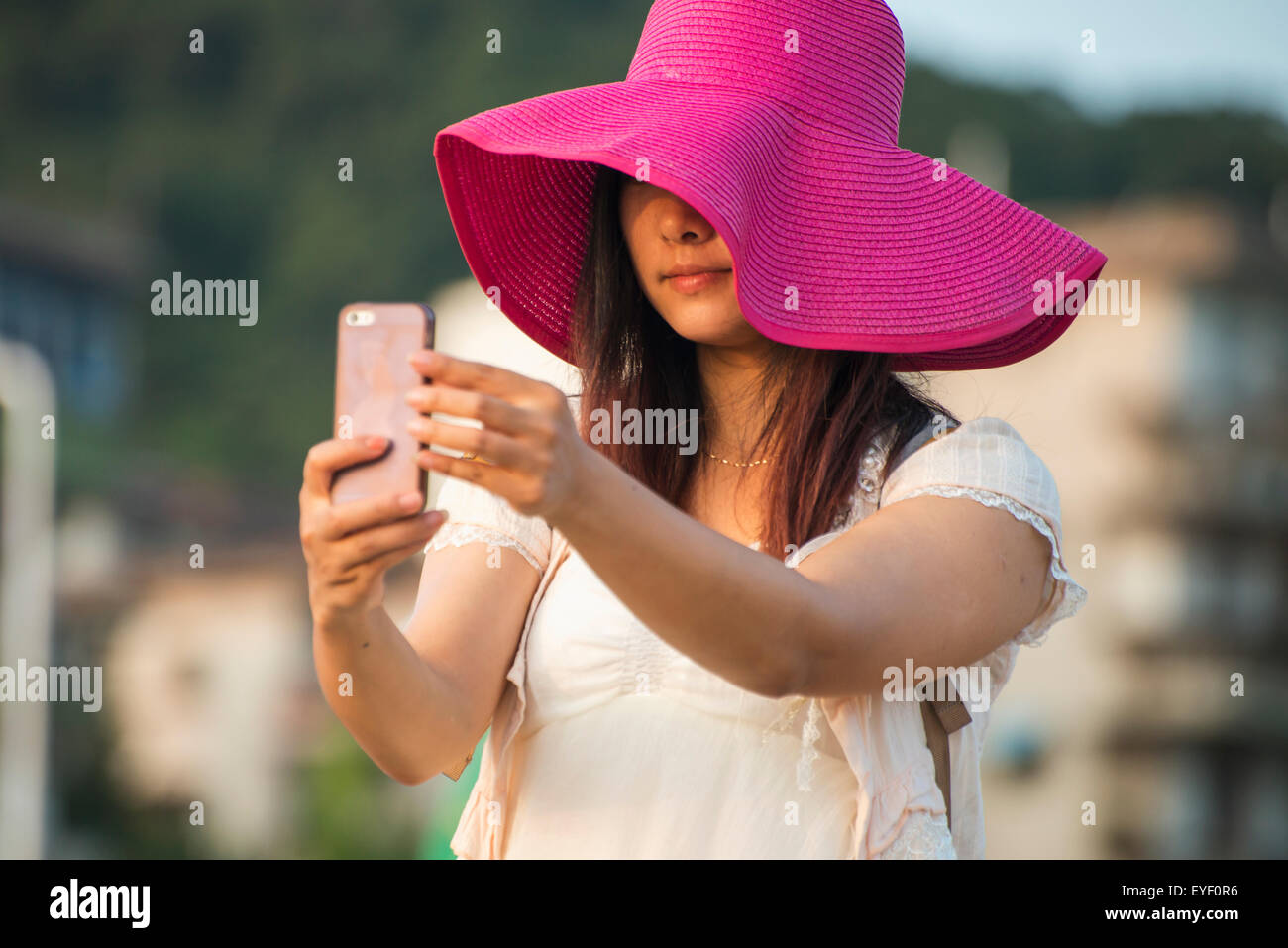 A young woman in a large brimmed pink hat takes a self-portrait with her cell phone; Xiamen, Fujian, China Stock Photo