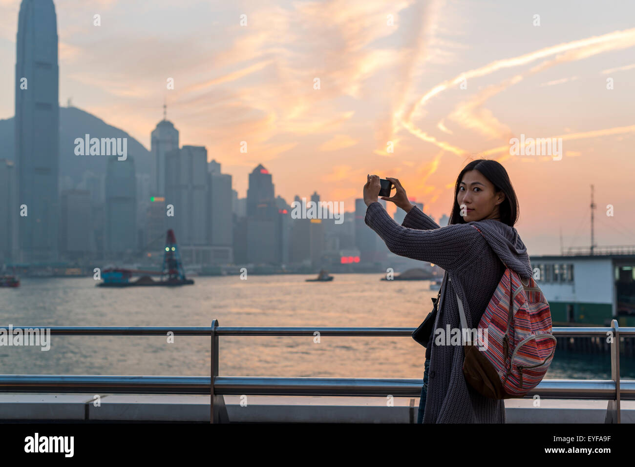 A young woman at the waterfront taking pictures of the skyline at sunset, Kowloon; Hong Kong, China Stock Photo