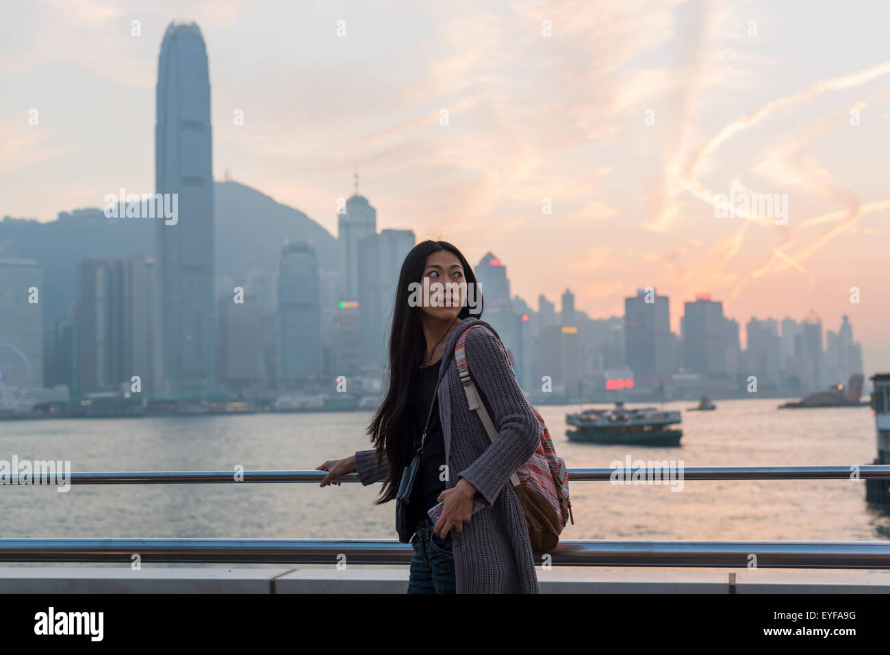 A young woman at the waterfront with a view of the skyline at sunset, Kowloon; Hong Kong, China Stock Photo