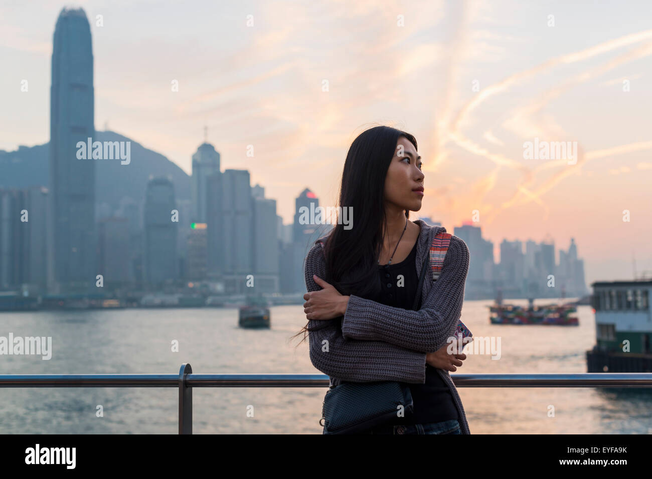 A young woman at the waterfront with a view of the skyline at sunset, Kowloon; Hong Kong, China Stock Photo