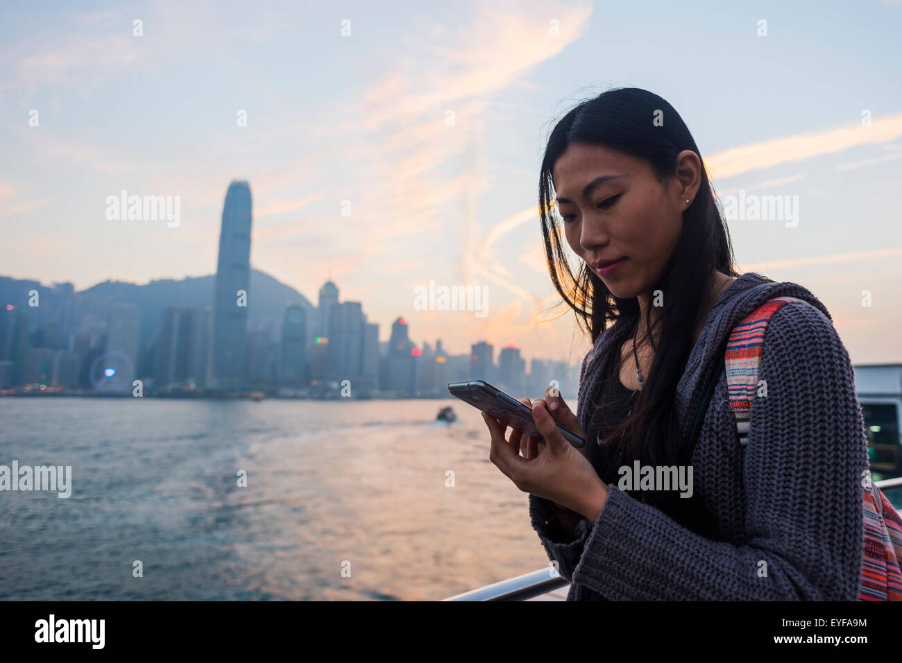 A young woman checks her cell phone at the waterfront with a view of the skyline at sunset, Kowloon; Hong Kong, China Stock Photo
