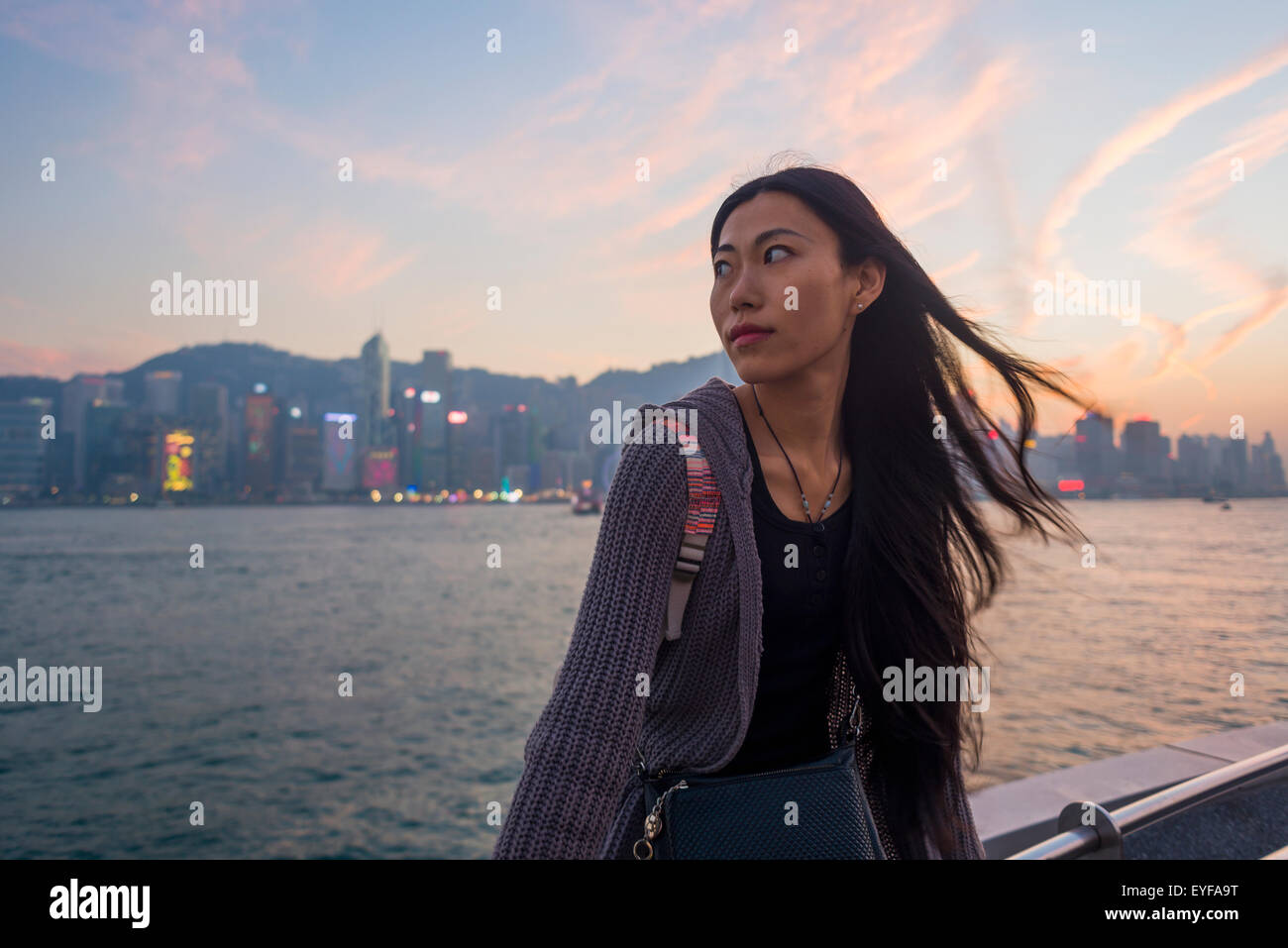 A young woman at the waterfront sitting on the railing at sunset with the skyline in the background, Kowloon; Hong Kong, China Stock Photo