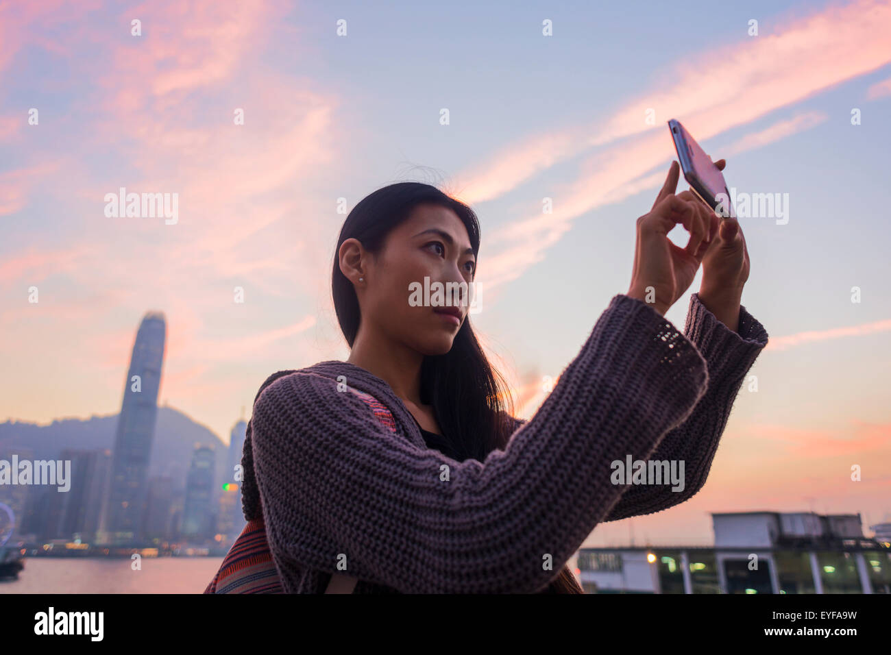 A young woman at the waterfront taking a picture at sunset with the skyline in the background, Kowloon; Hong Kong, China Stock Photo