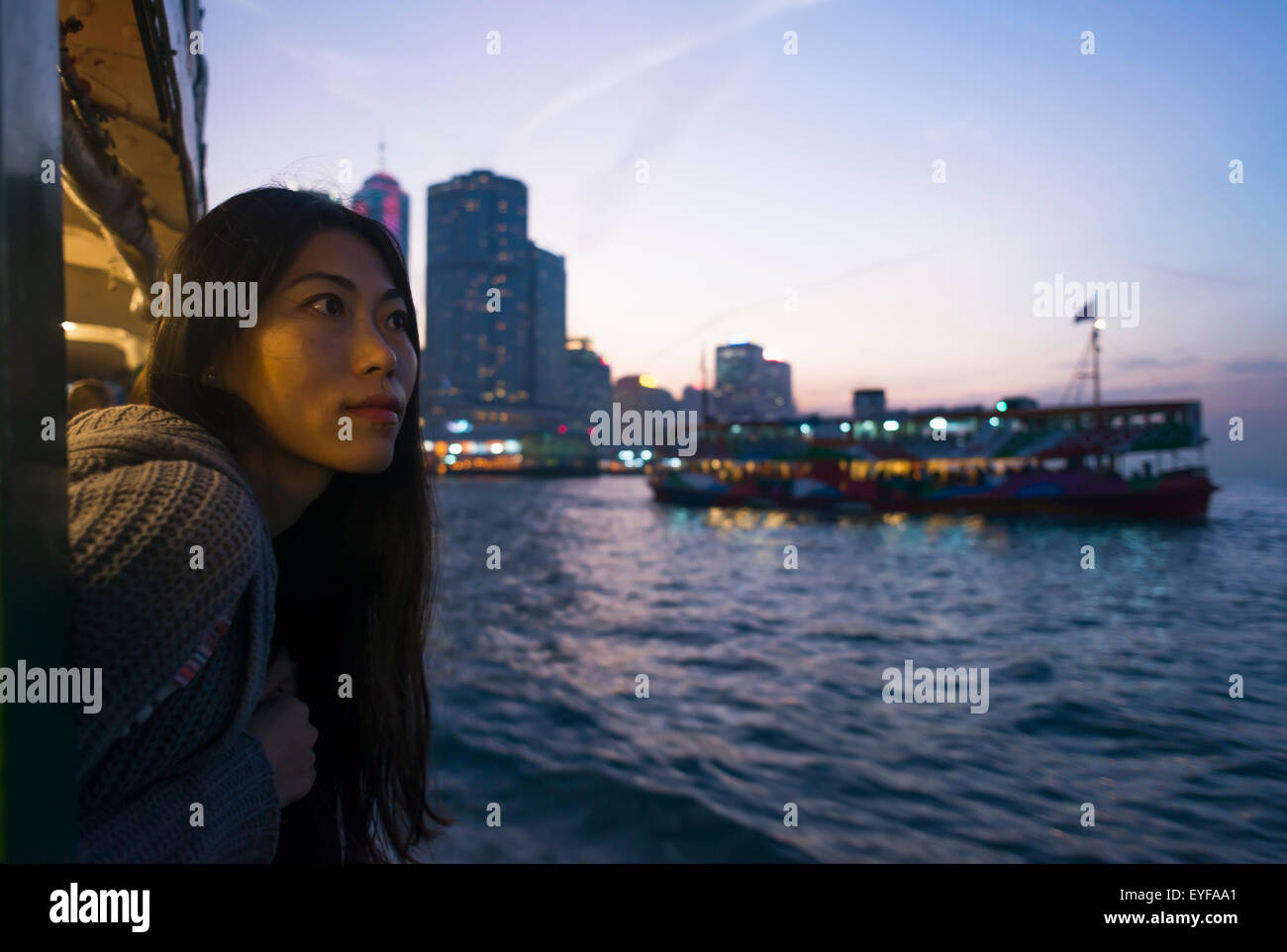 A young woman at the waterfront at sunset with a a boat and skyline in the background, Kowloon; Hong Kong, China Stock Photo