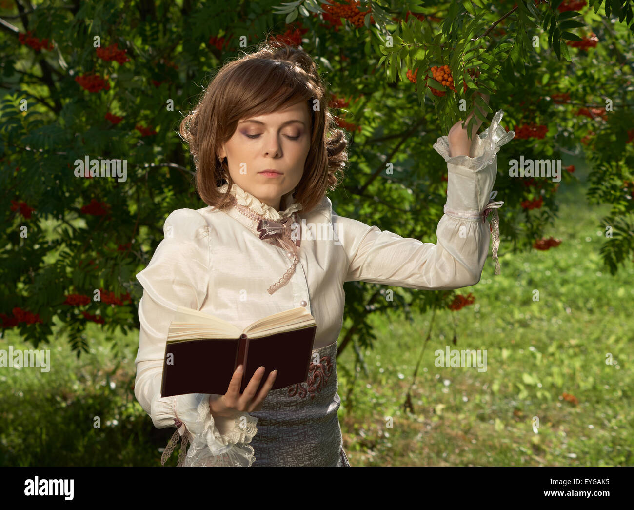 Beautiful girl in old style clothing touches rowan berries and reads book Stock Photo