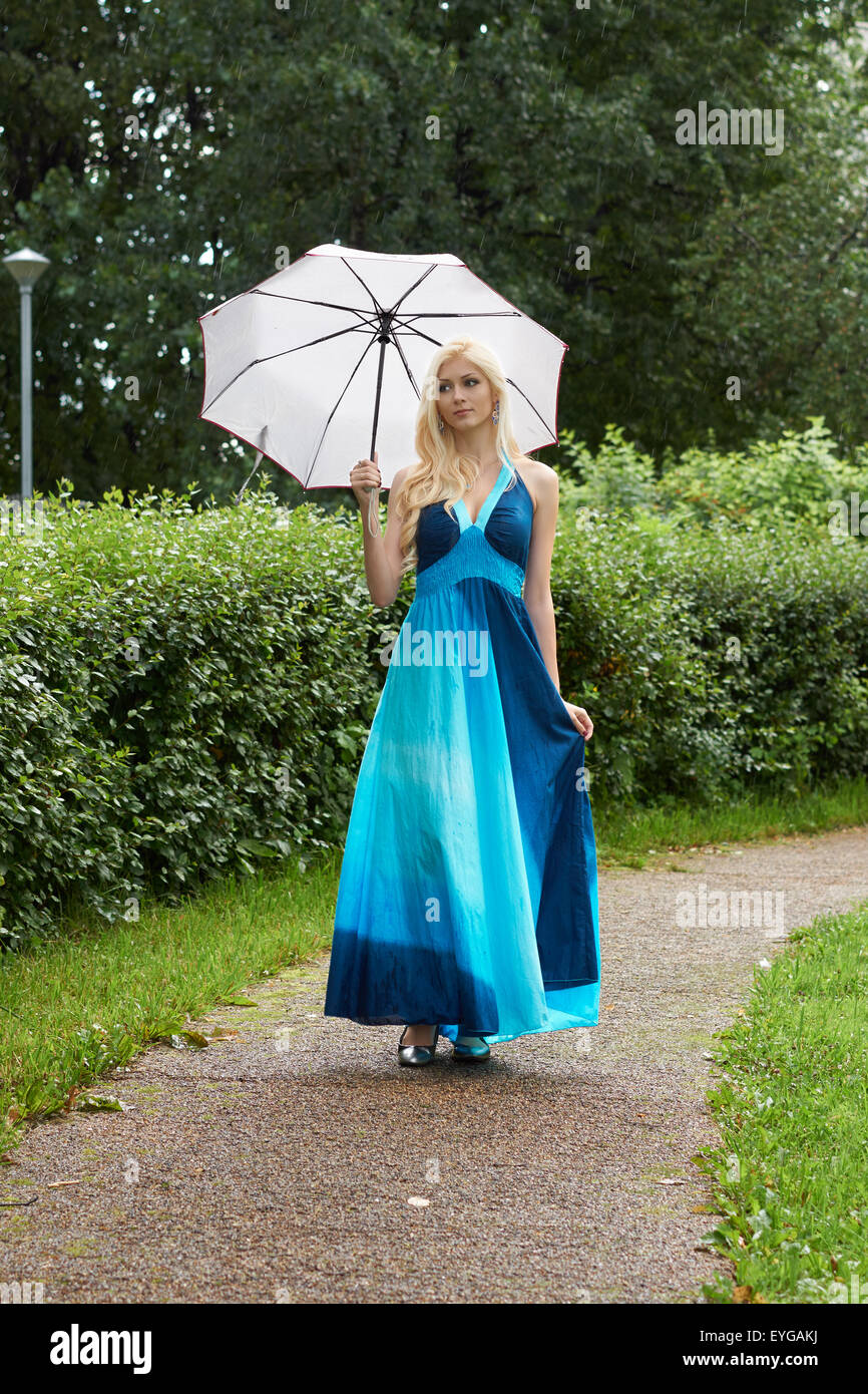 Beautiful blond girl with umbrella stands on walkway in the park under rain Stock Photo