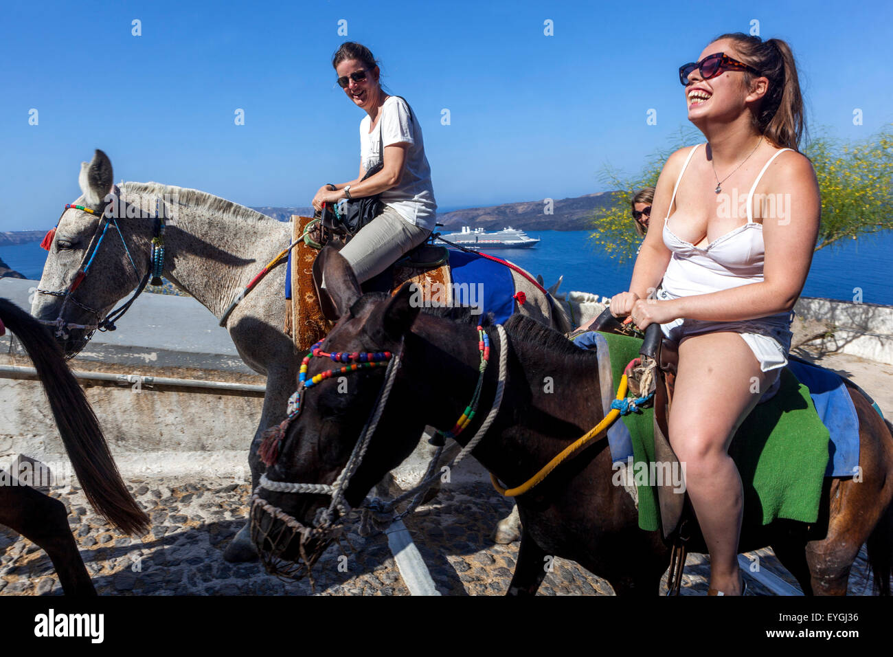 Greece tourists enjoying life, People, Two young women riding donkeys and laughing. Road linking the port to the town of Thira Santorini Greece Stock Photo