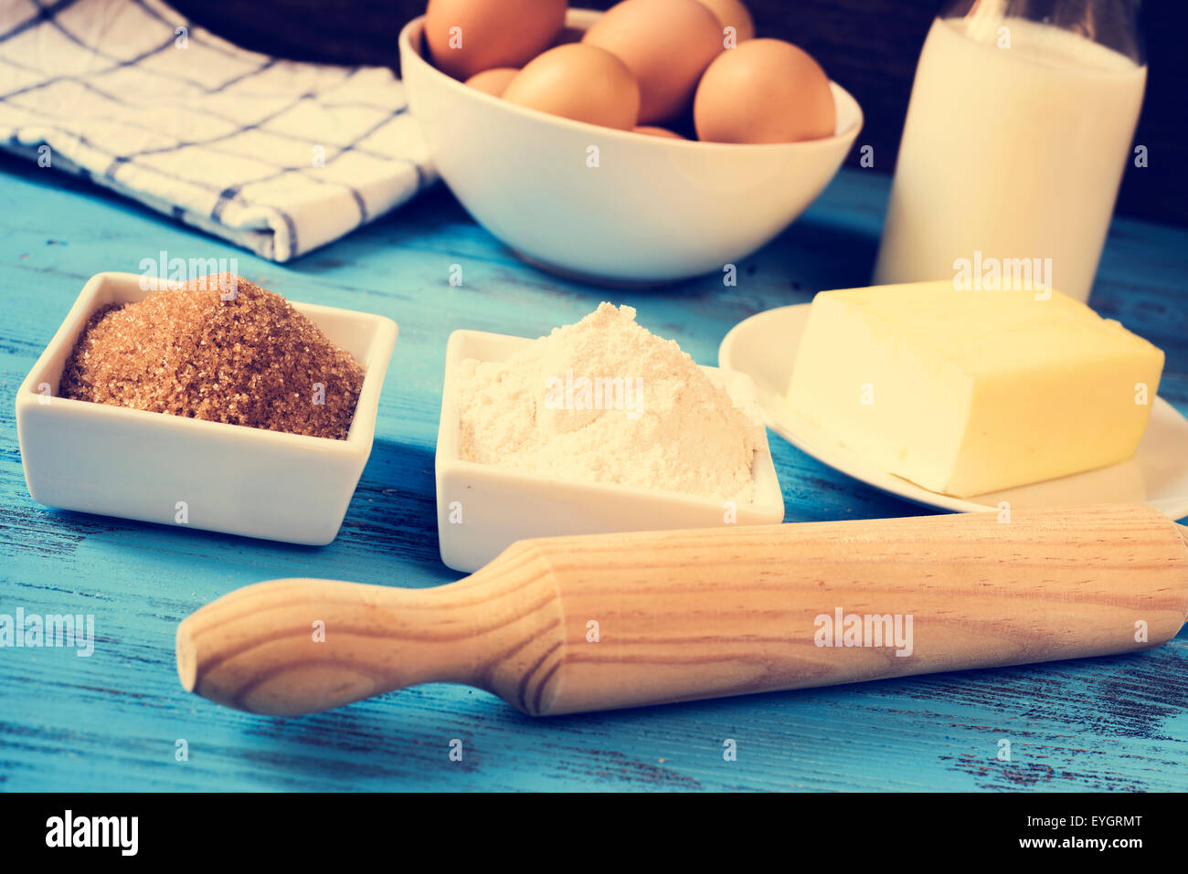some ingredients for preparing cookies or a cake, such as milk, eggs, flour, butter and brown sugar on a blue rustic wooden surf Stock Photo