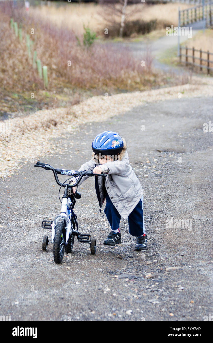 Boy struggling to push bicycle with stabilizers, up rough track in the