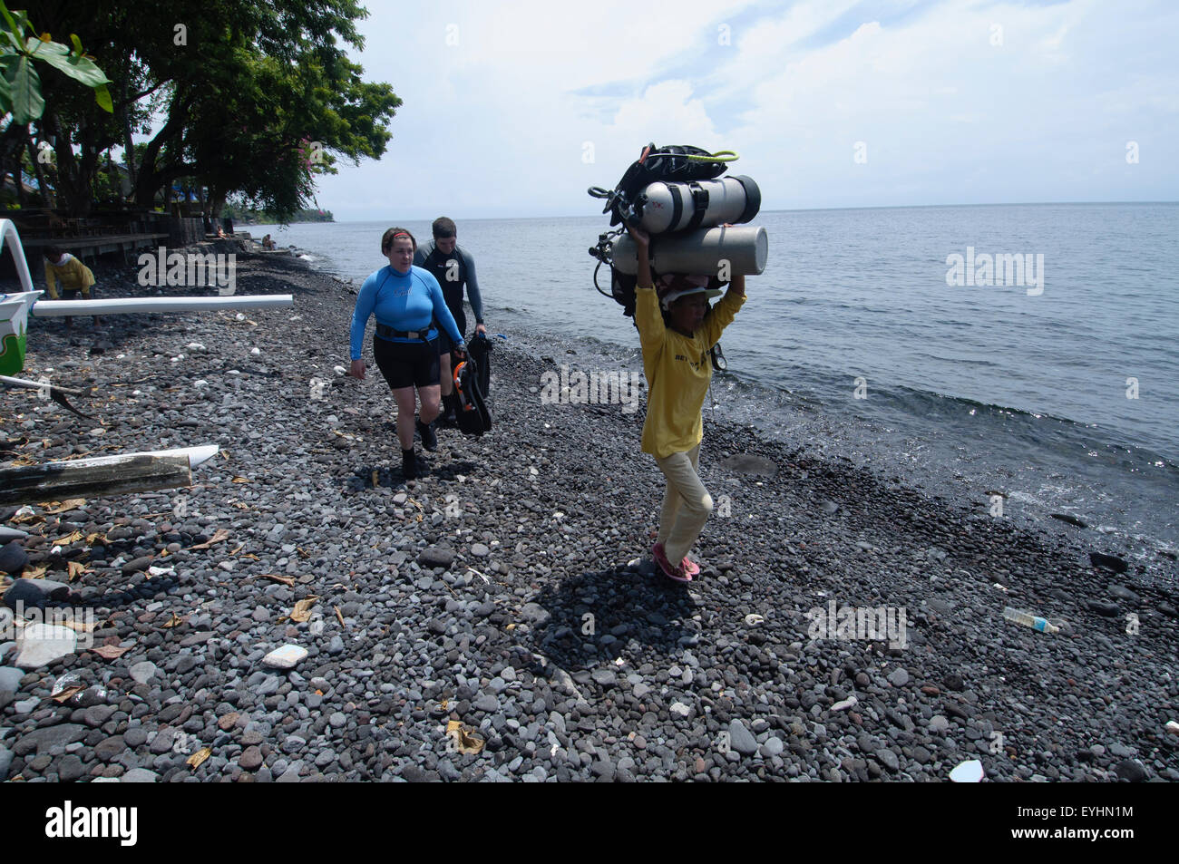A female porter carries two fully loaded scuba tanks on her head along a rocky beach while the divers follow along, Tulamben Stock Photo