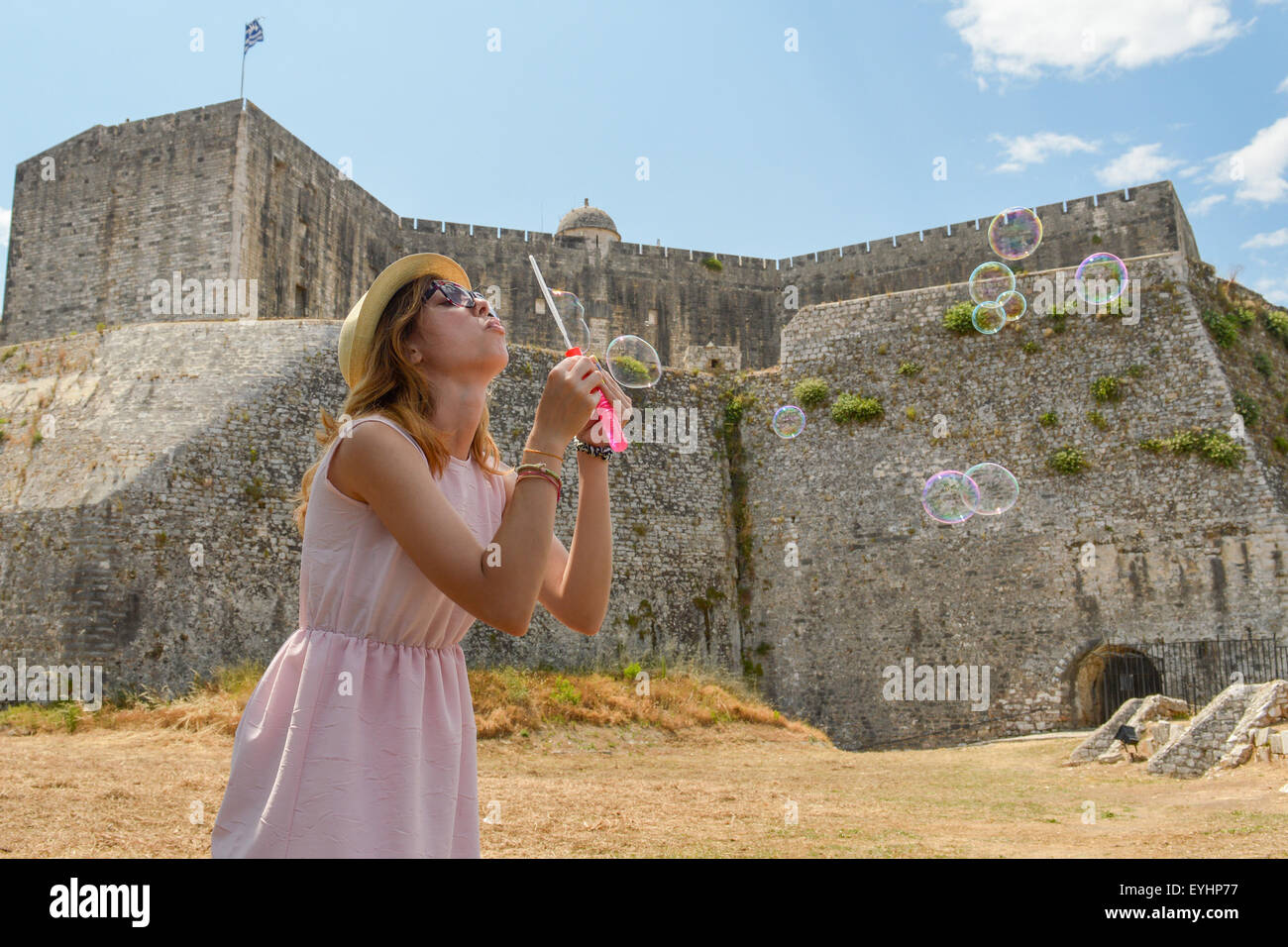 Young girl blowing soap bubbles at the fortress wearing straw hat and pink dress Stock Photo