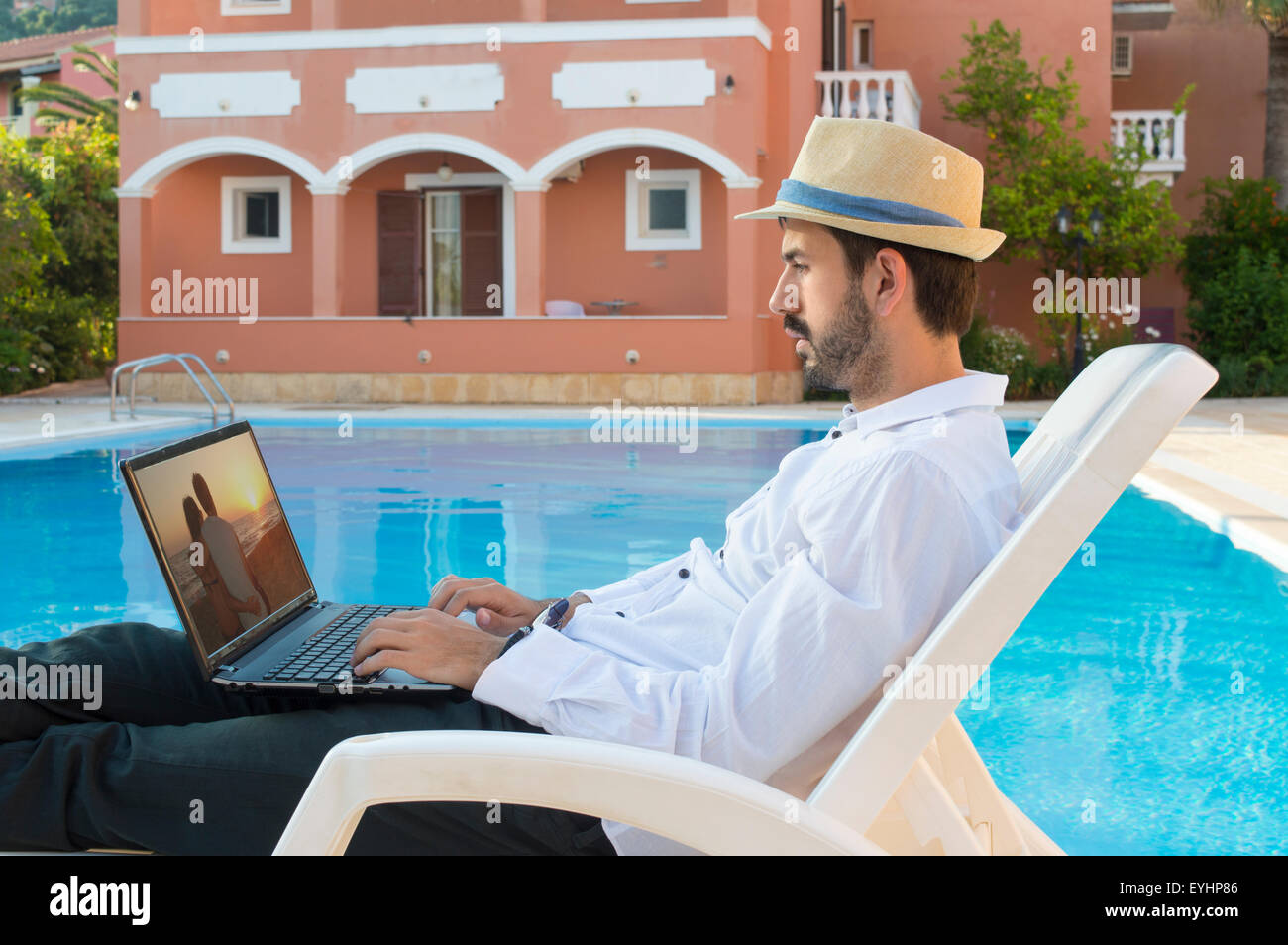 Young bussines man on pause while working on his lap top by the pool while on vacation  wearing straw hat Stock Photo