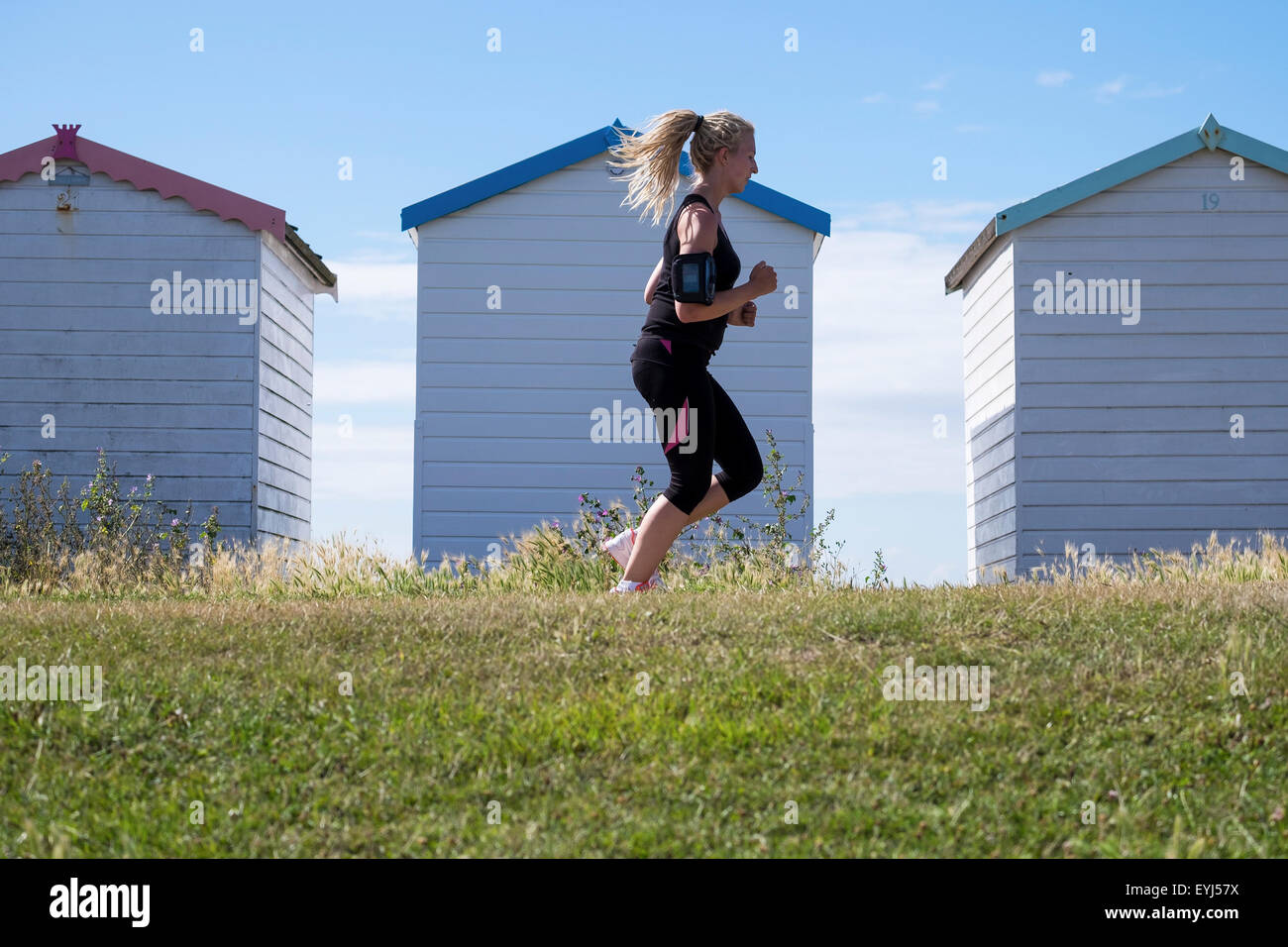 Young woman with tied back hair blonde dreadlocks wearing short leggings running along seafront past row of beach huts Stock Photo