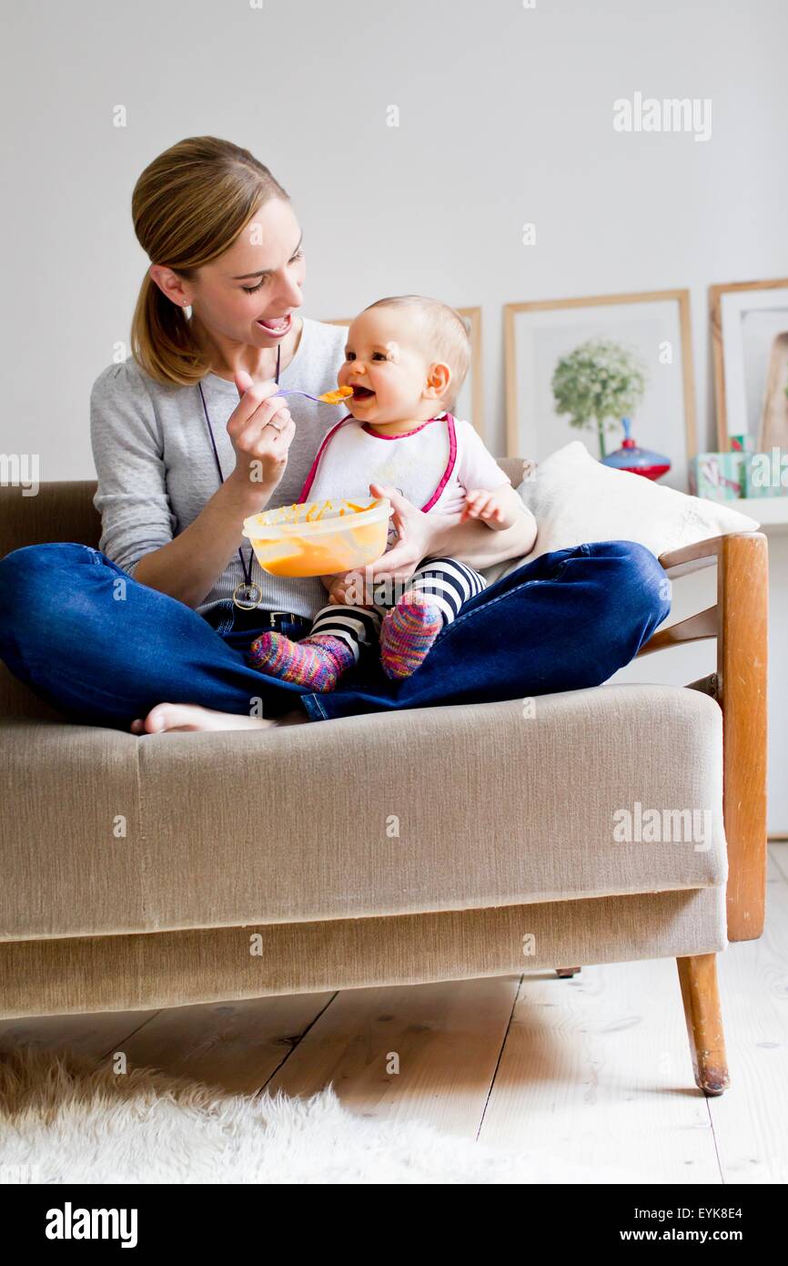 Mother sitting on sofa, feeding baby girl Stock Photo