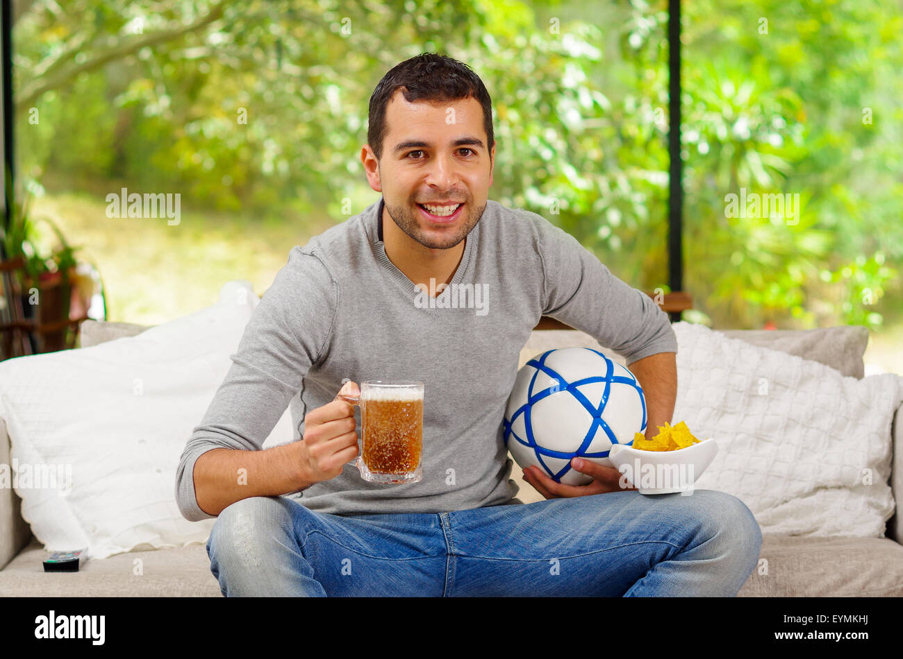 Hispanic man wearing denim jeans with grey sweater sitting in sofa enthusiastically watching tv holding beer and football Stock Photo