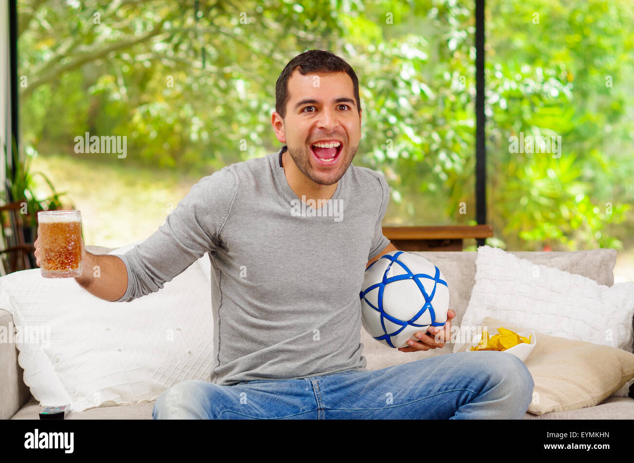 Hispanic man wearing denim jeans with grey sweater sitting in sofa enthusiastically cheerful facial expression watching tv holding beer and football Stock Photo