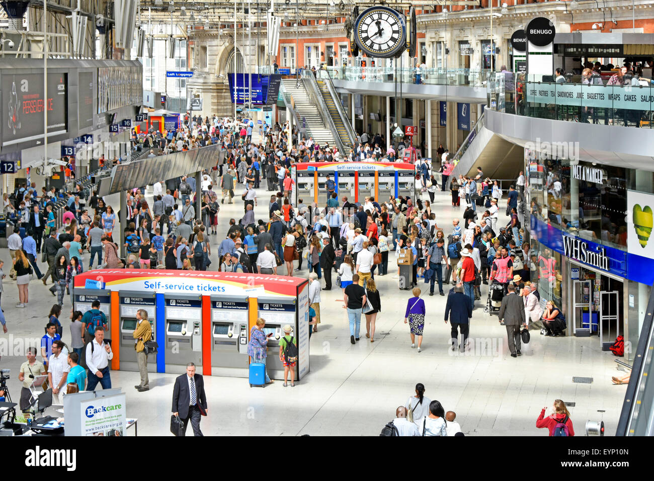 Commuters on Waterloo train station concourse with station clock self service train ticket machines retail shops & balconies London England UK Stock Photo