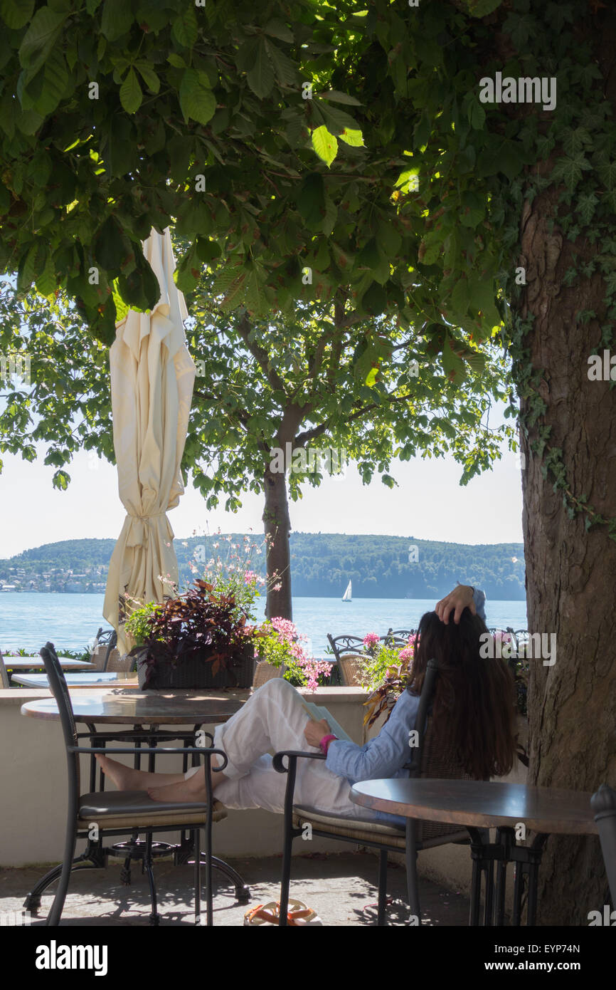 A young woman reading a book in the shade in a cafe restaurant by the shores of a lake Stock Photo
