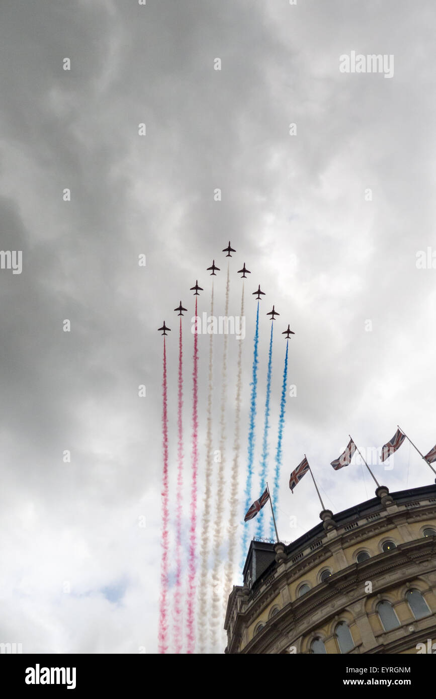 Trafalgar Square, London, Engand. The Red Arrows fly past streaming red white and blue over a building with Union Jack  flags. Stock Photo