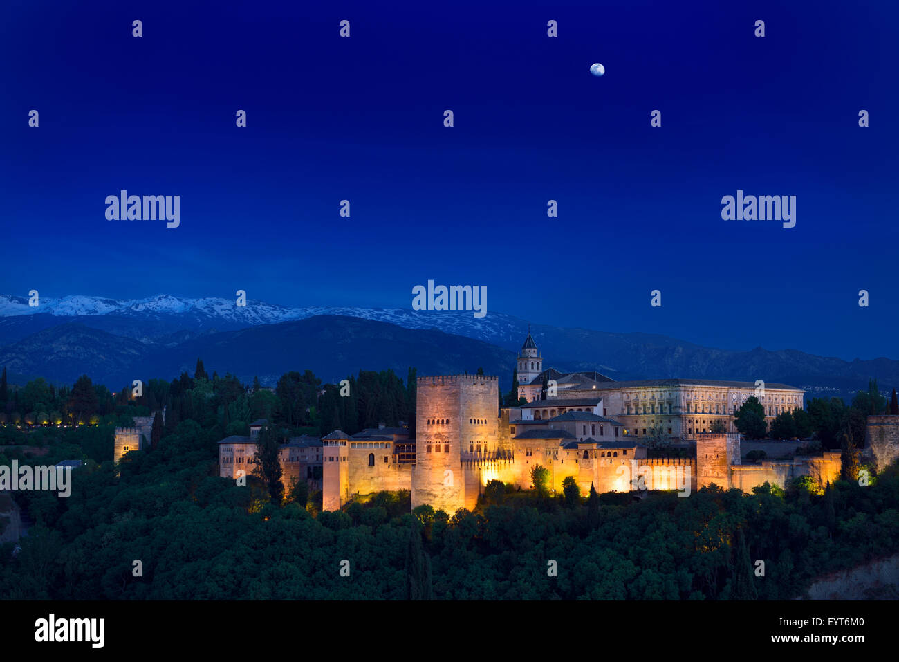 Moonrise over lit hilltop Alhambra Palace fortress complex at twilight Granada with snow capped Sierra Nevada Mountains Spain Stock Photo
