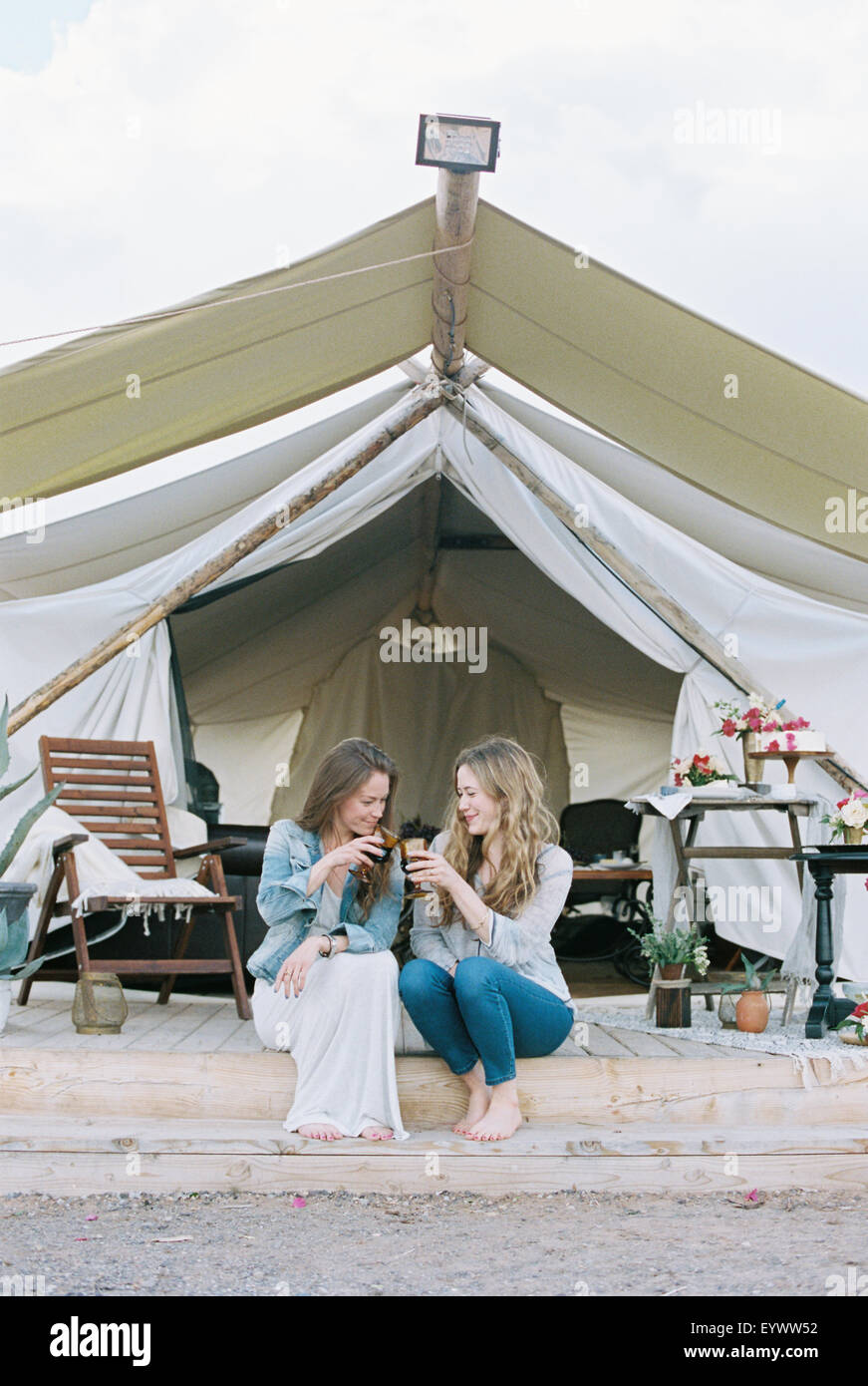 Two smiling women sitting outside a large tent laughing and having a glass of wine. Stock Photo