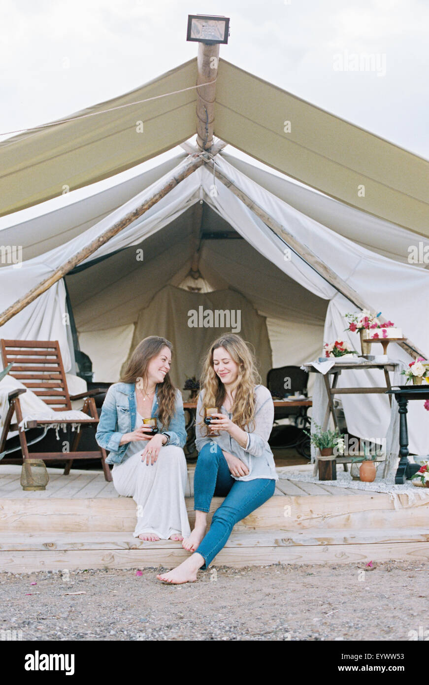Two smiling women sitting outside a large tent laughing and having a glass of wine. Stock Photo