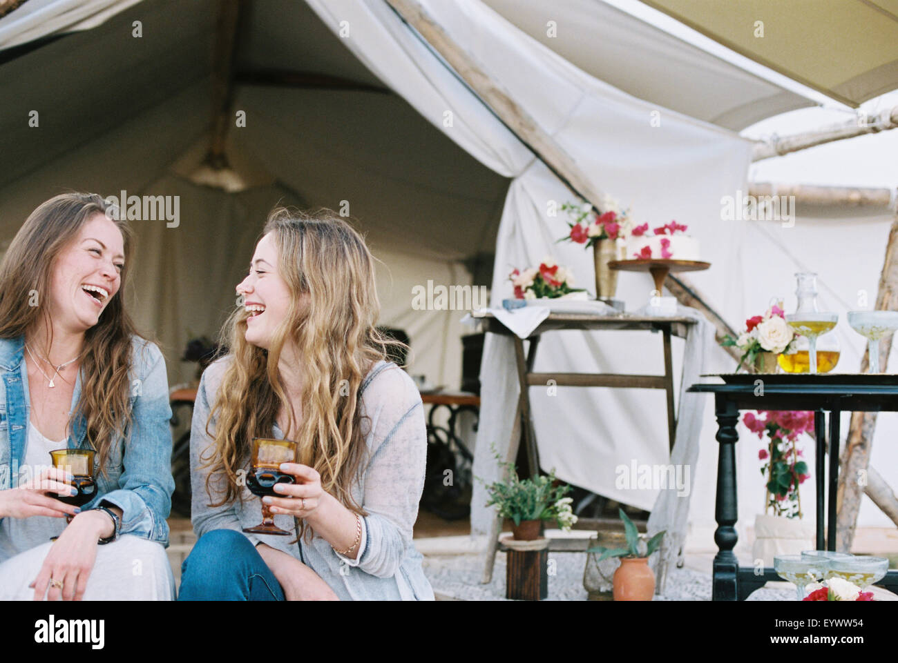 Two smiling women sitting outside a tent in a desert, enjoying a glass of wine. Stock Photo