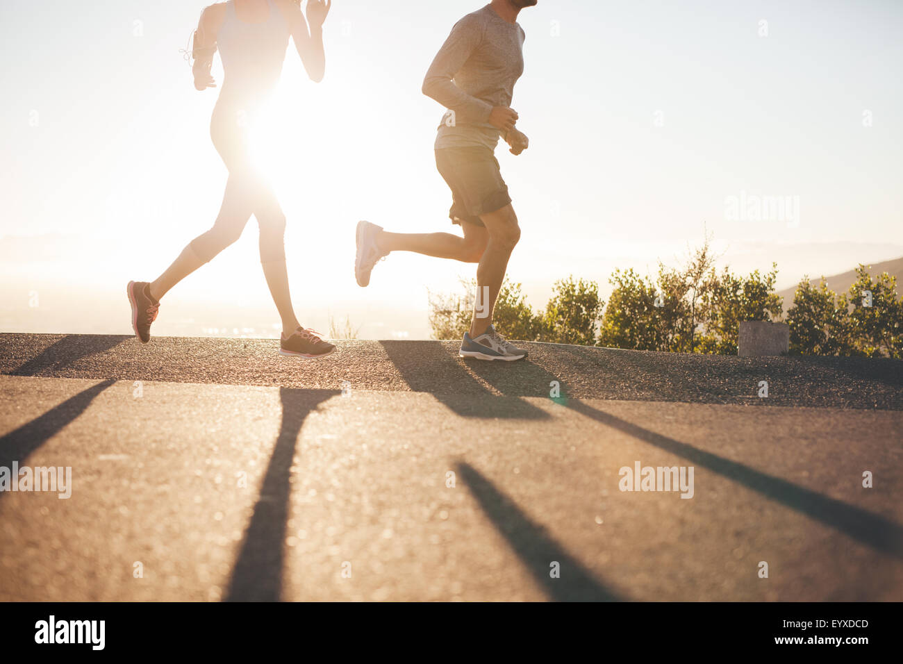 Low angle view of two people running on country road at sunrise. Cropped shot of young man and woman jogging in morning, with br Stock Photo