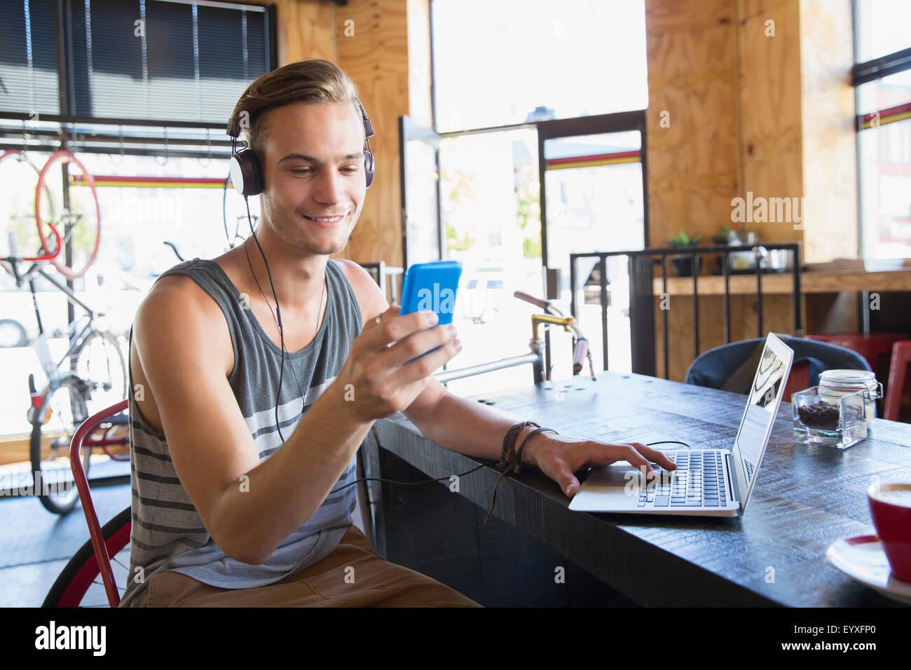 Smiling young man with headphones texting with cell phone at laptop in cafe Stock Photo